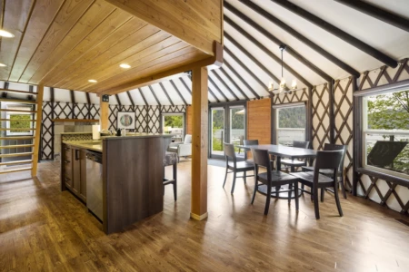 This is a spacious Moutcha Bay Resort Yurt, well-lit kitchen with a dining area, featuring wooden floors, beams, and a vaulted ceiling. Large windows offer outdoor views.